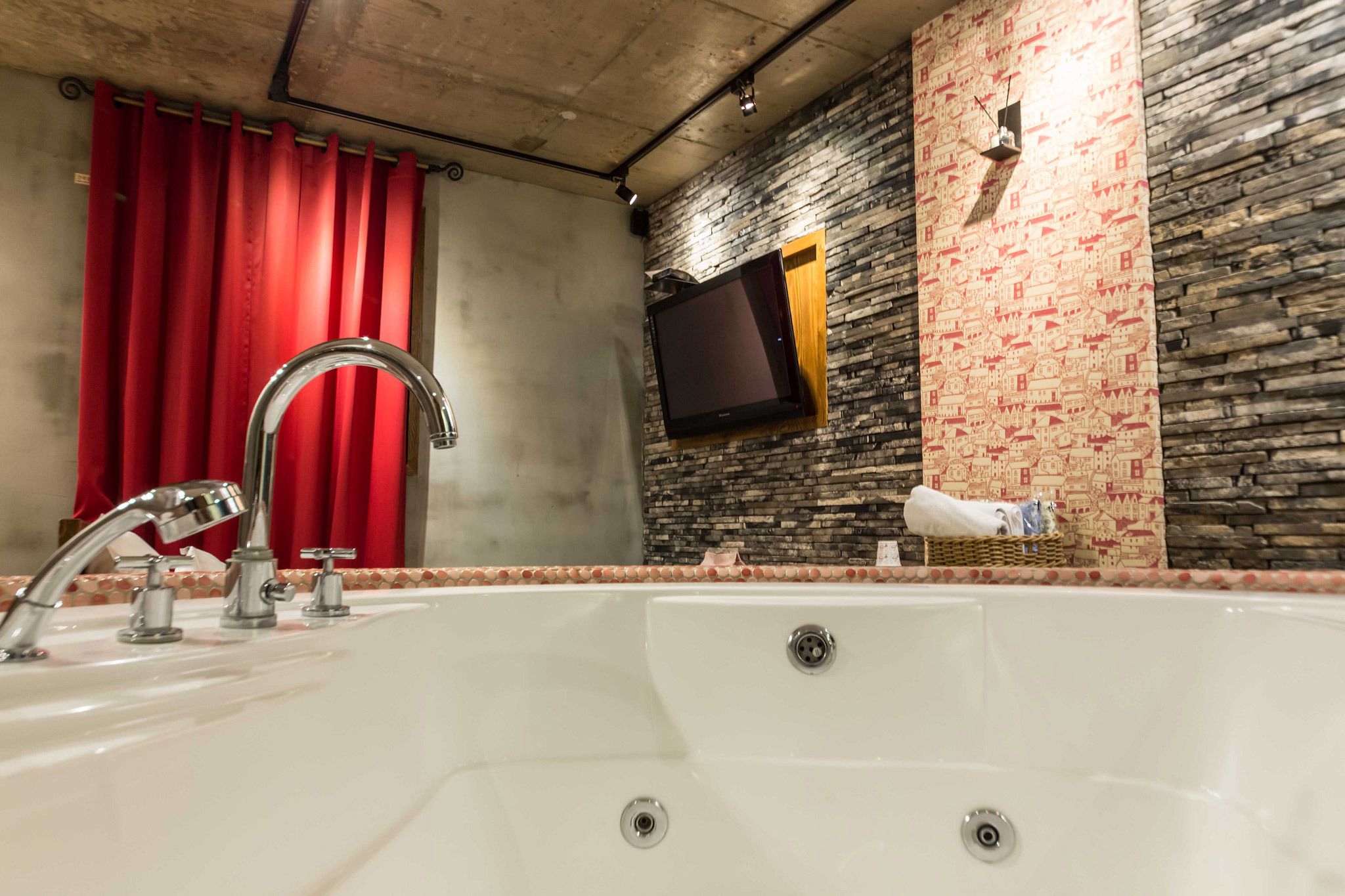Een badkamer met een groot bad, een rood gordijn, een tv aan de muur en een wand met een steenstructuur.