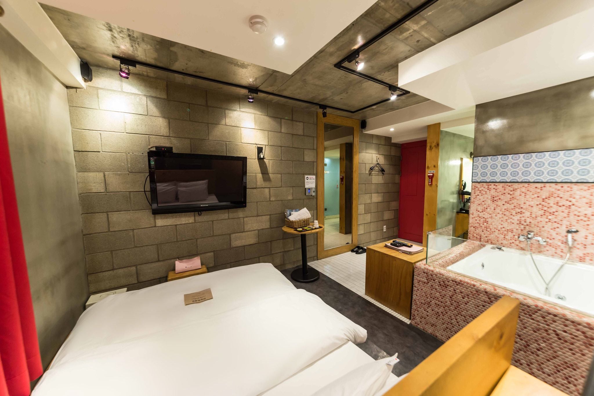 A modern hotel room with a large bed, a small table, a TV, a bathroom with a bathtub, and a mirror.
