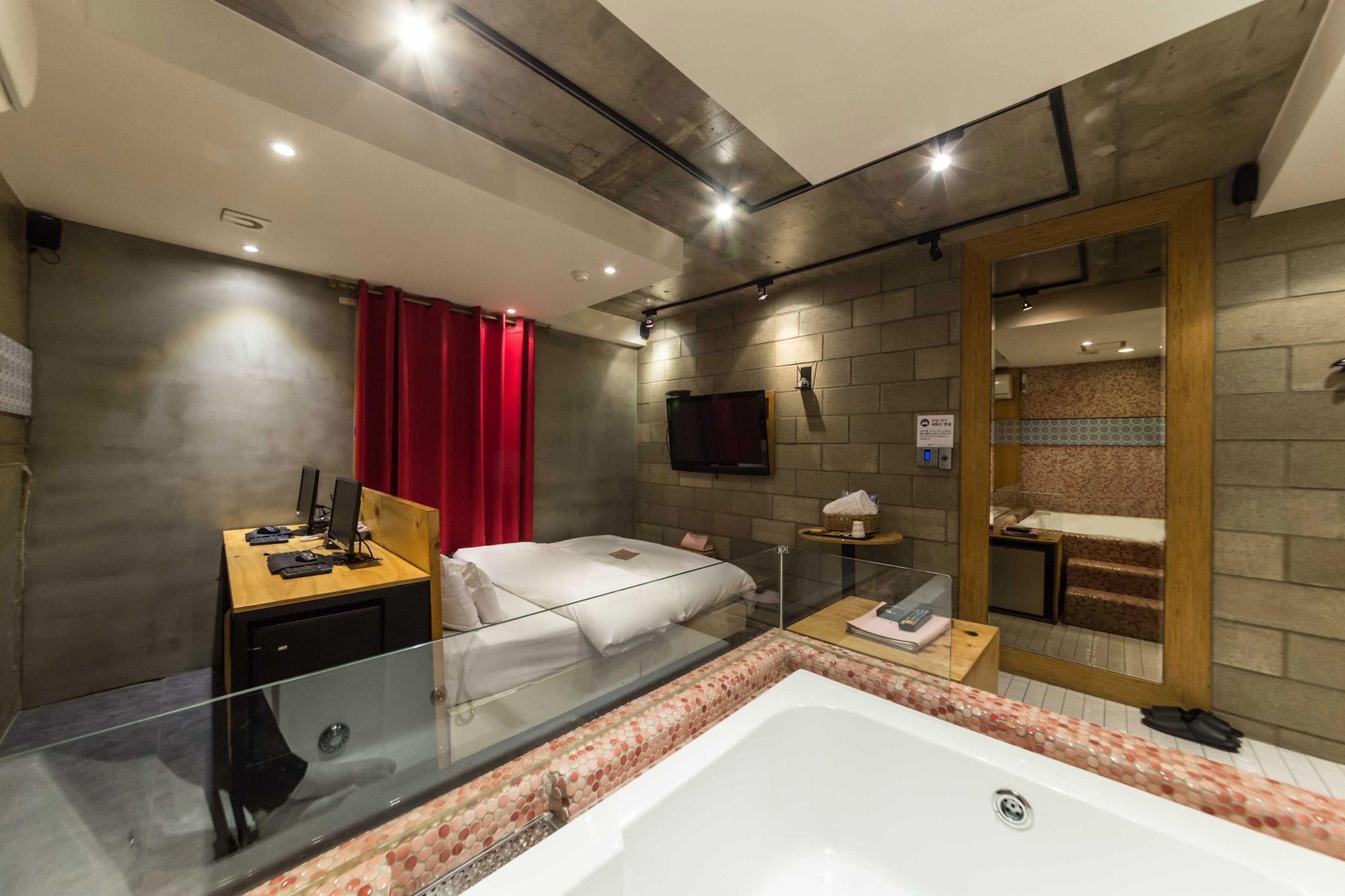 A modern hotel room with a glass-enclosed balcony, a large bathtub, a bed, a small desk with a computer, and a television mounted on the wall.