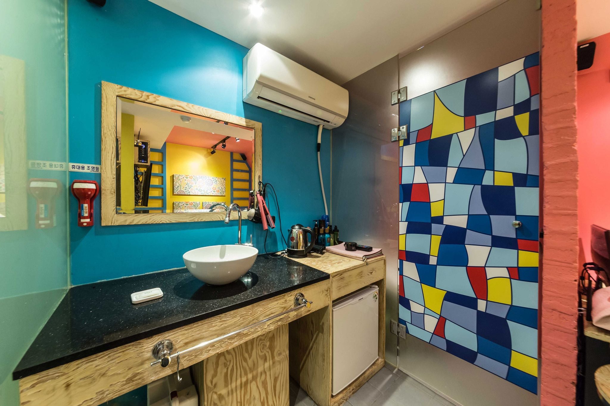 Un baño con pared azul, lavamanos blanco, espejo y un diseño abstracto y colorido en la pared.