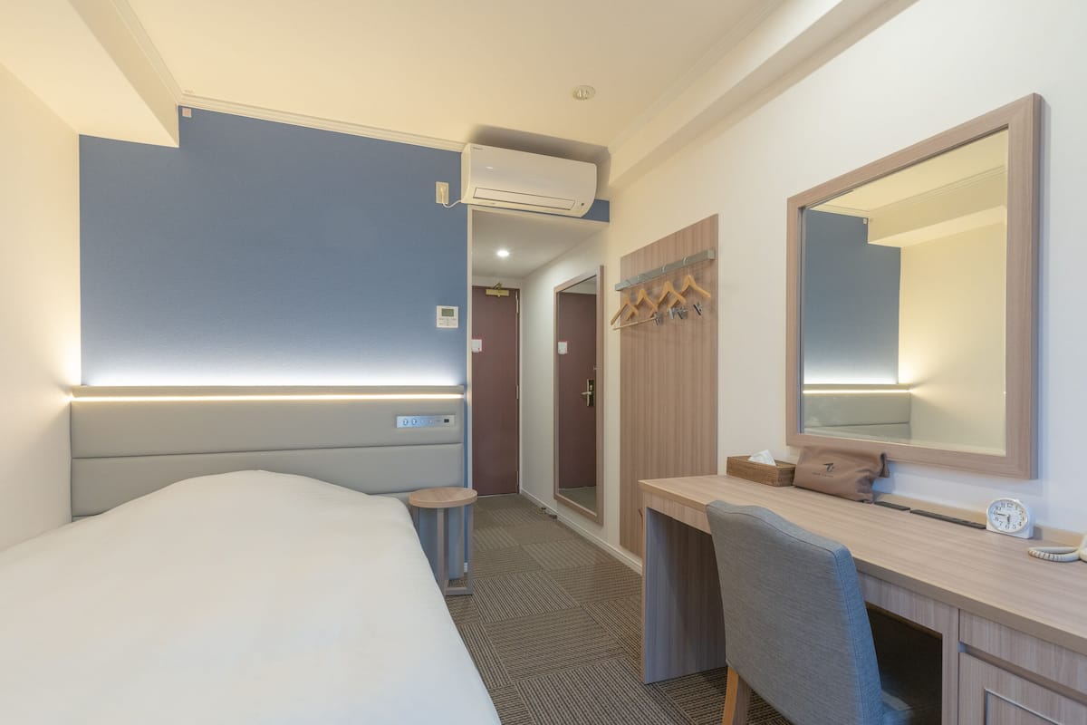 A hotel room with a bed, a desk, a chair, a mirror, a clock, and a hanging coat rack.