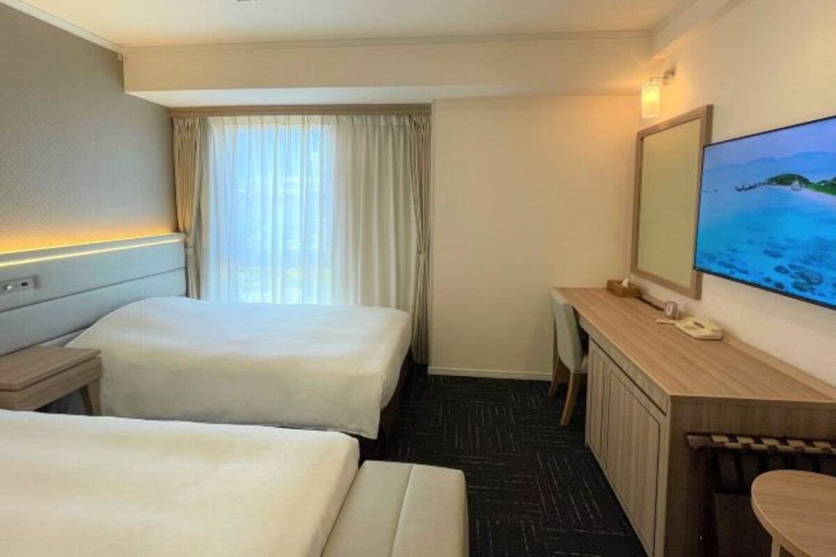 A hotel room with two beds, a desk, a chair, a TV, and a window with curtains.