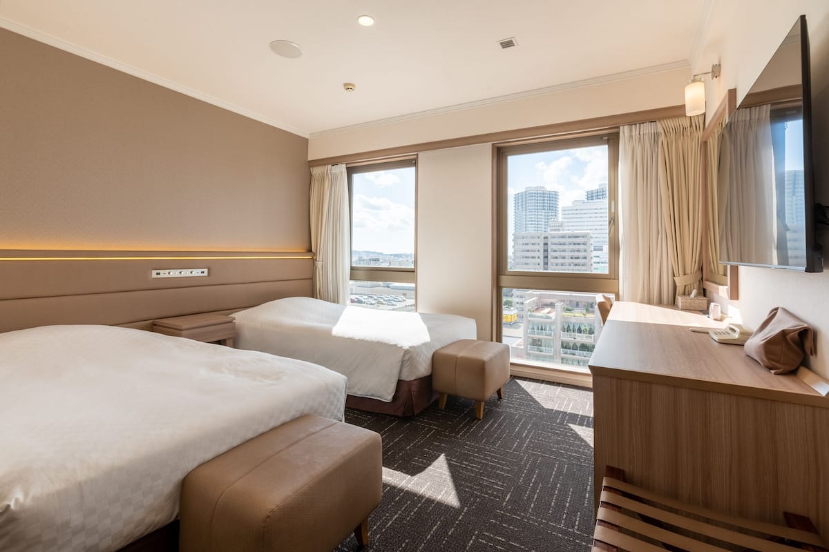 A hotel room with two beds, a desk, a chair, a large window with a city view, and a flat-screen TV.