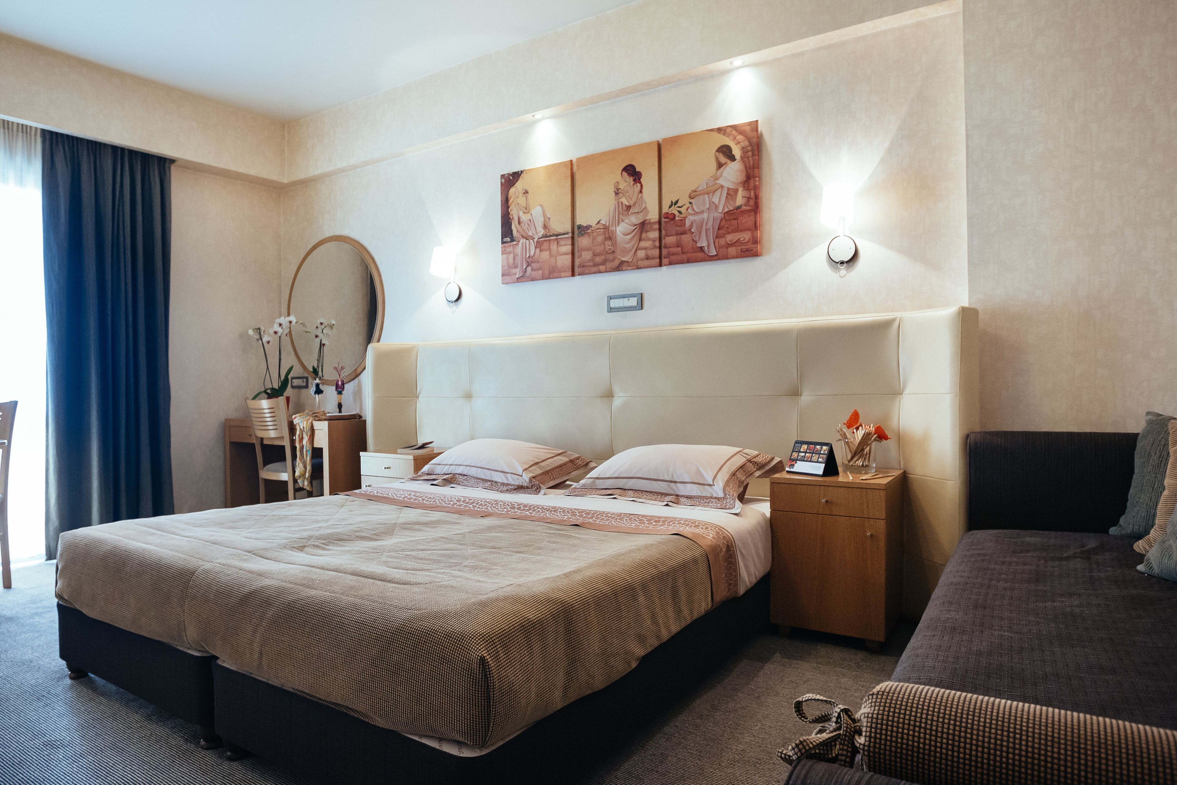 A hotel room with a large bed, a bedside table, a nightstand, a chair, a desk, a mirror, and a wall-mounted artwork.