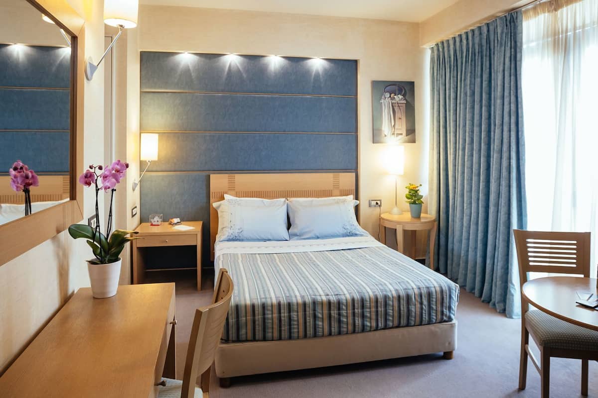 A hotel room with a bed, bedside tables, a desk, a chair, a mirror, and a potted plant.
