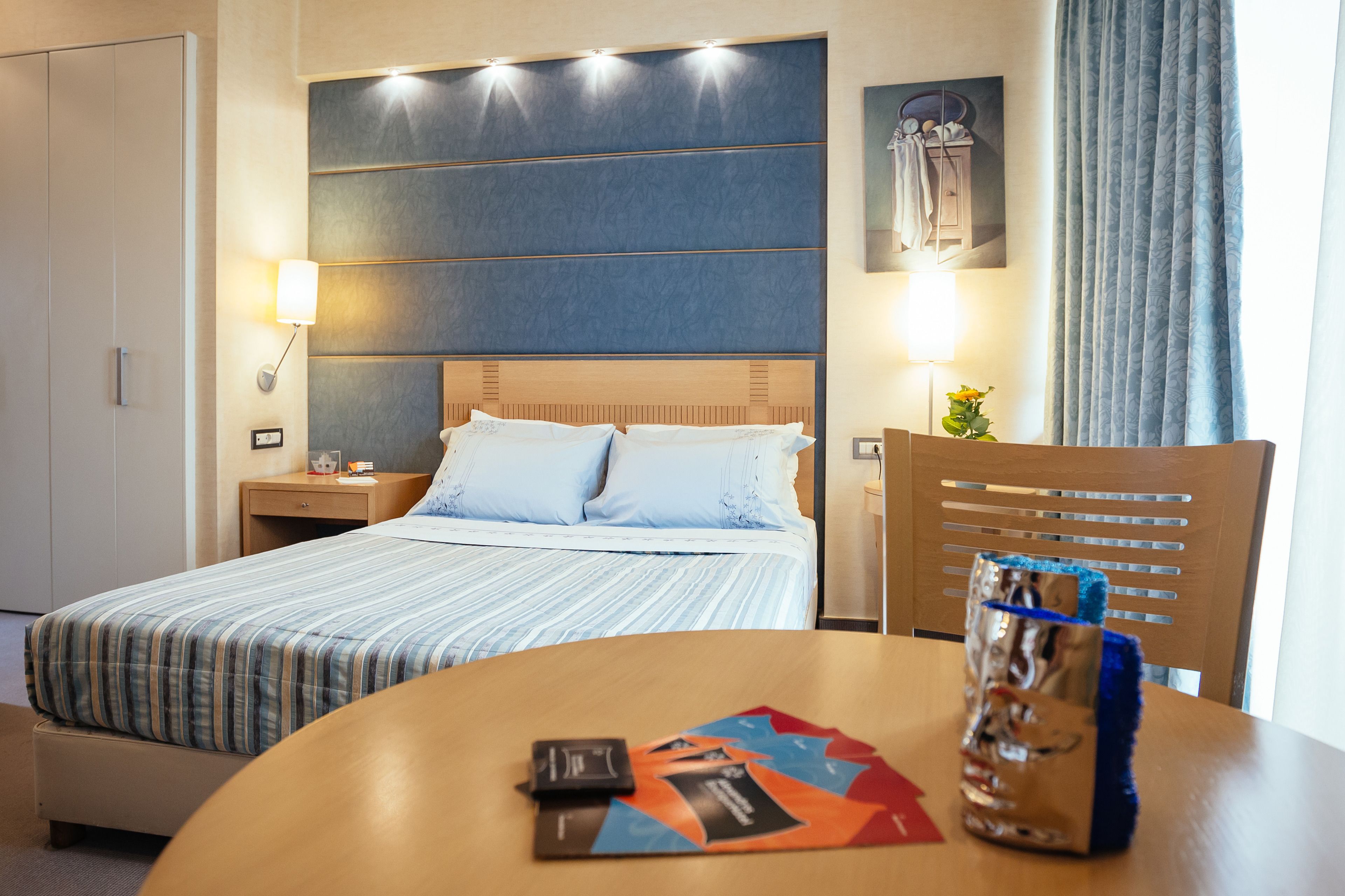 A hotel room with a bed, bedside tables, a chair, and a table with a card and a glass.