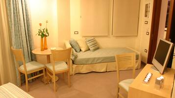 Una habitación pequeña y acogedora con una cama, un escritorio con computadora, dos sillas, una mesita y un florero con flores.