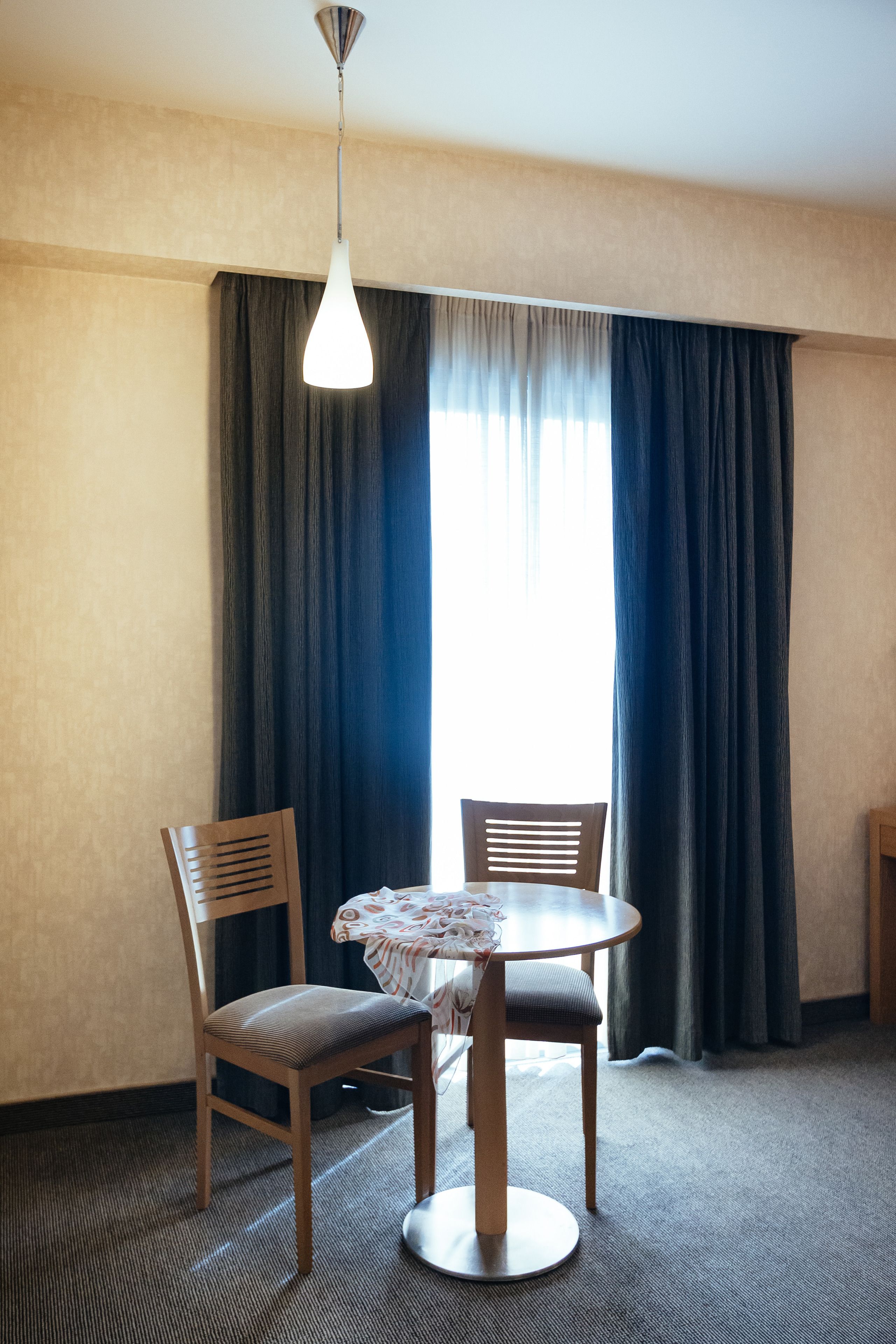 Quarto de hotel com mesa redonda, duas cadeiras, luminária pendente e uma janela grande com cortinas escuras.