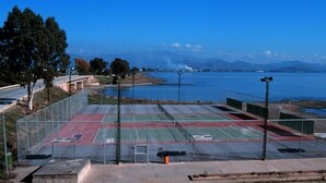 An outdoor tennis court with a metal fence, a metal railing, and a view of a body of water and mountains in the background. - Alexandros Boutique Hotel (Argos-Mykines)