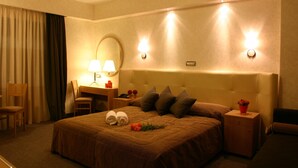 A hotel room with a large bed, a desk with a lamp, a chair, a nightstand with a red flower vase, and a wall-mounted mirror. - Alexandros Boutique Hotel (Argos-Mykines)