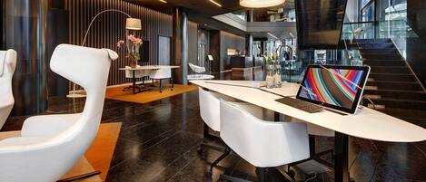 Un espacio de oficina moderno con un escritorio blanco, una computadora portátil y una silla blanca. Se aprecia un ventanal grande al fondo, y una escalera a la derecha.