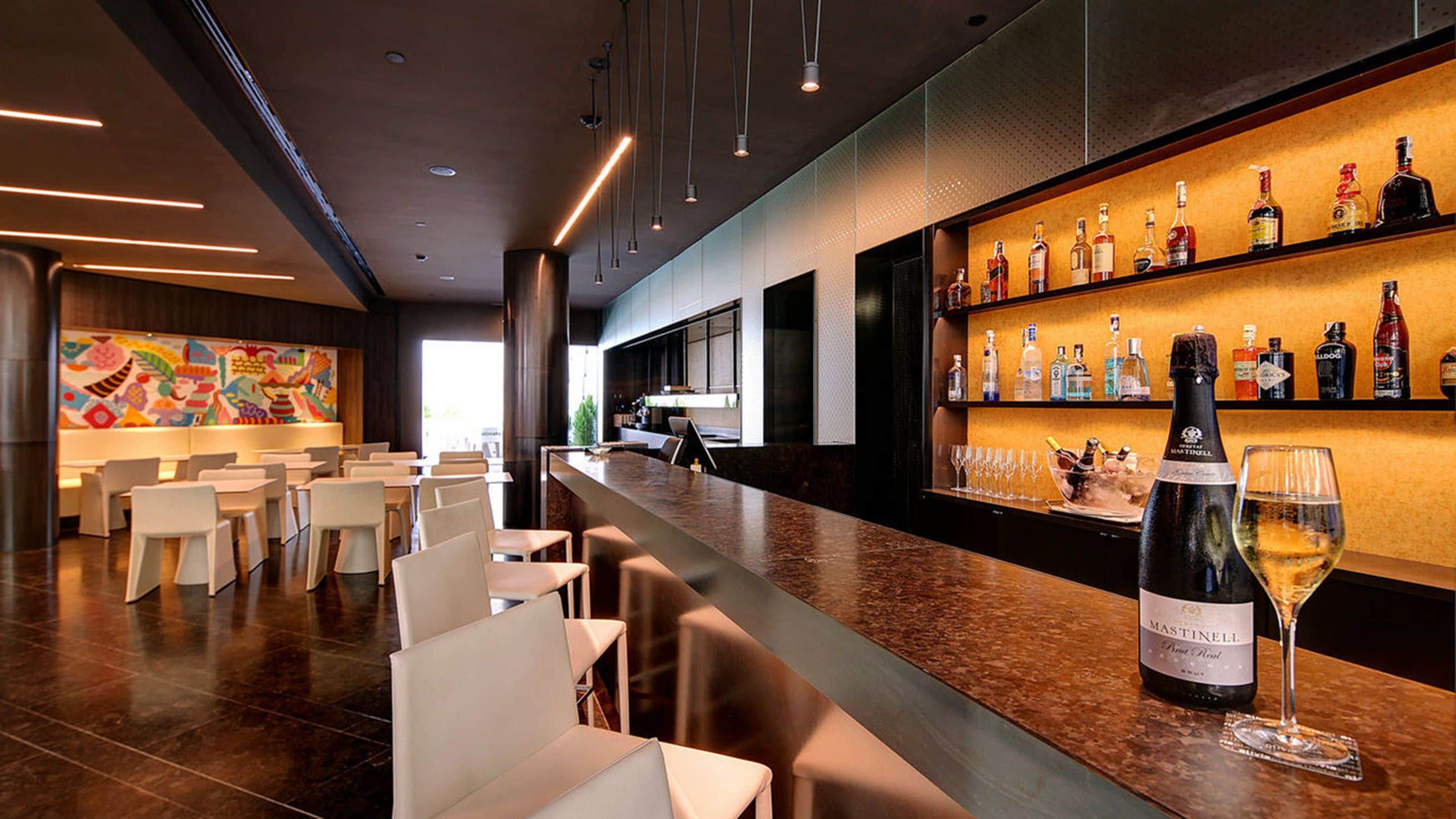 A modern bar area with a long counter, white chairs, and a variety of liquor bottles.
