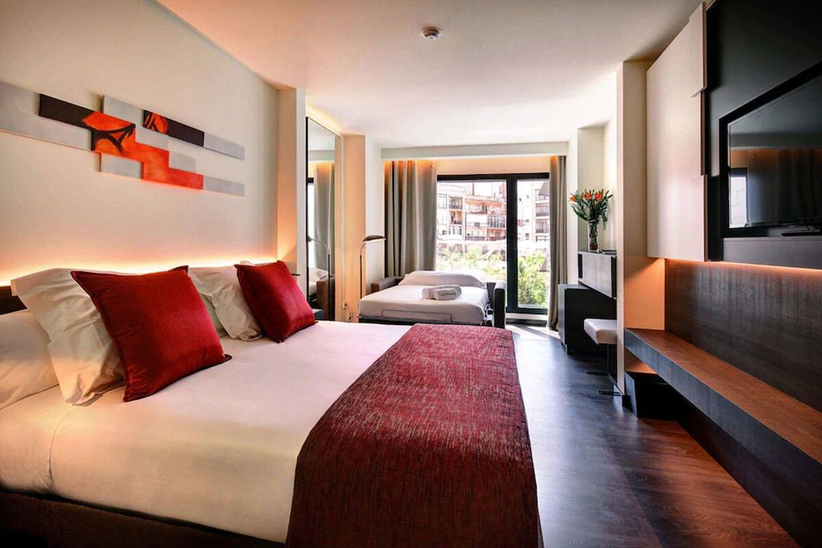 A modern hotel room with a large bed, a TV, and a balcony with a view of the city.