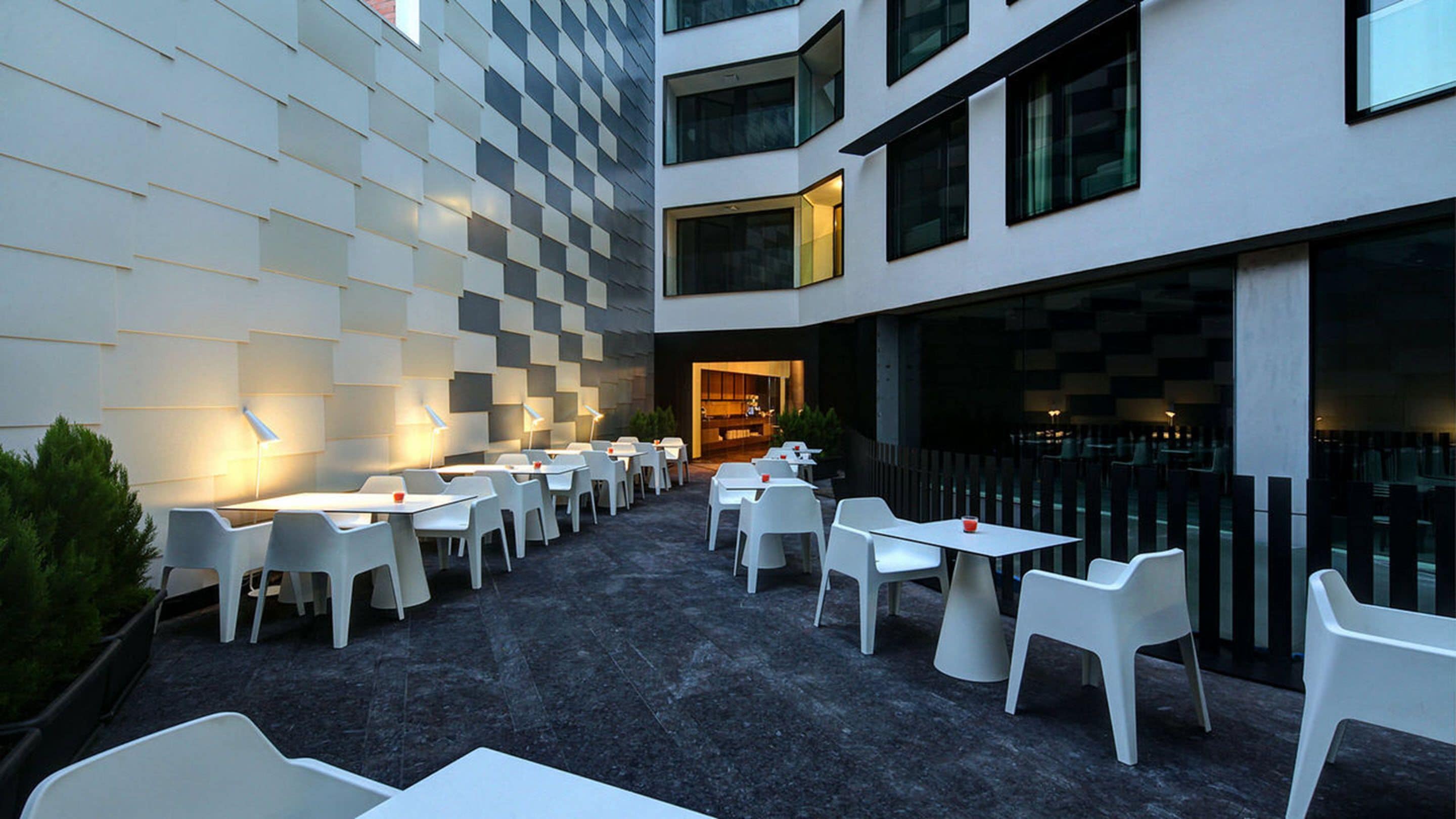 An outdoor dining area with white tables and chairs, a checkered wall, and a modern building facade.