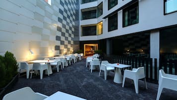 Un espace de restauration en plein air avec des tables et des chaises blanches, un mur à carreaux et la façade d’un bâtiment moderne.