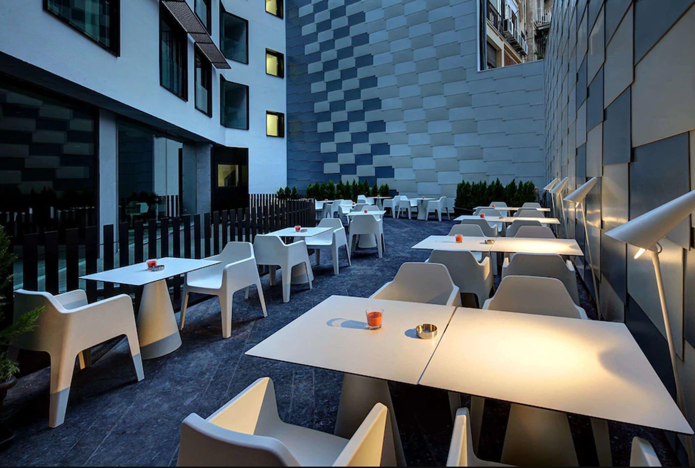 Un área de comedor al aire libre con mesas y sillas blancas, un muro a cuadros y una fachada de edificio moderna.