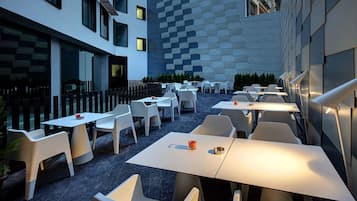 An outdoor dining area with white tables and chairs, a checkered wall, and a modern building facade.