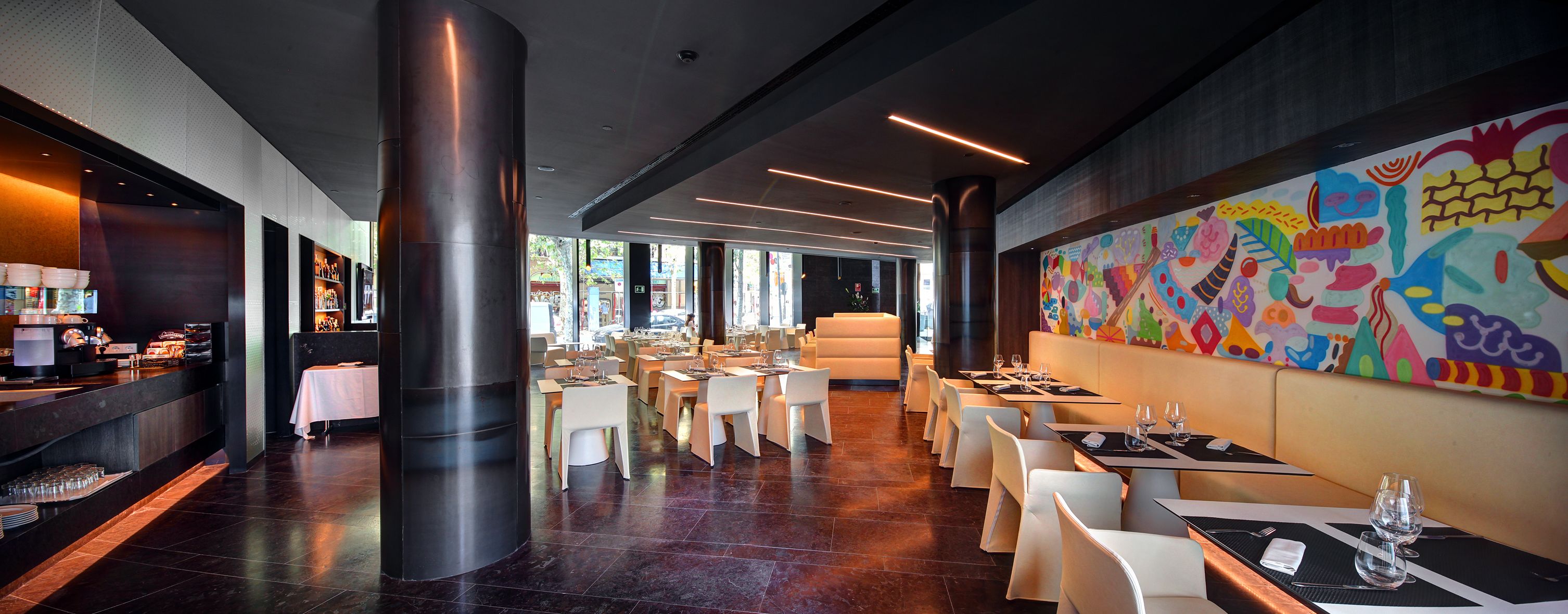 A modern restaurant with a central column, white tables, and a colorful mural on the wall.