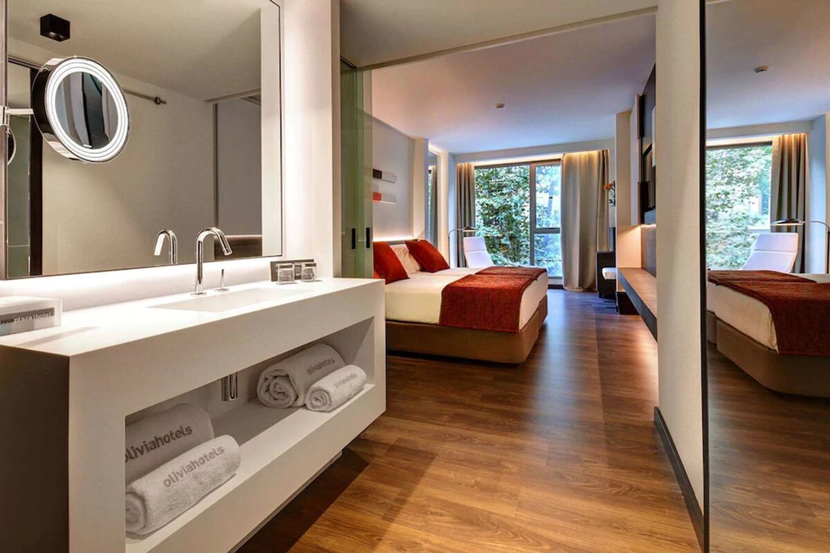 A modern hotel room with a large mirror, a vanity with a sink, a bed with red pillows, and a view of greenery through the windows.