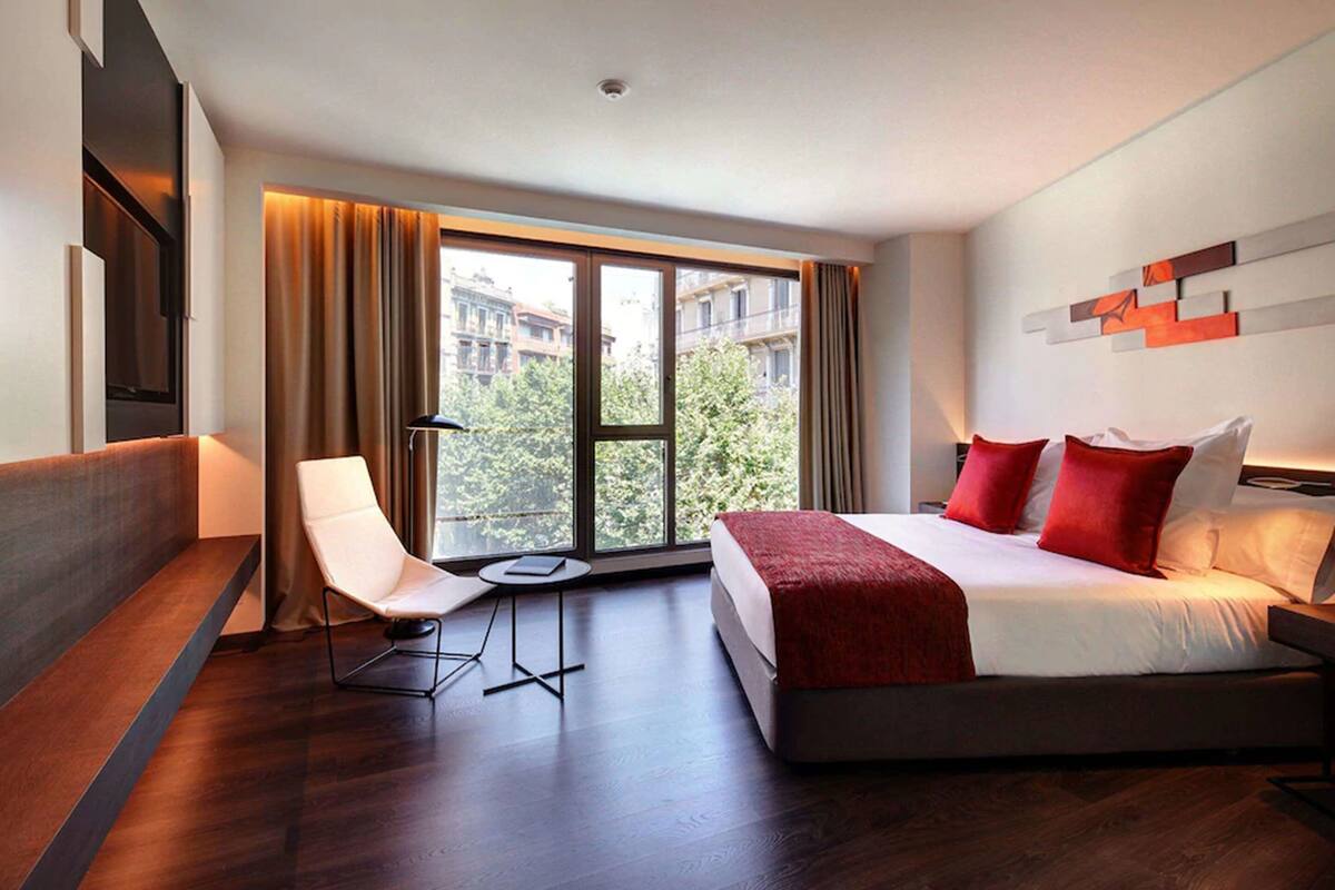 A modern hotel room with a large bed, a chair, a small table, and a view of buildings through large windows.