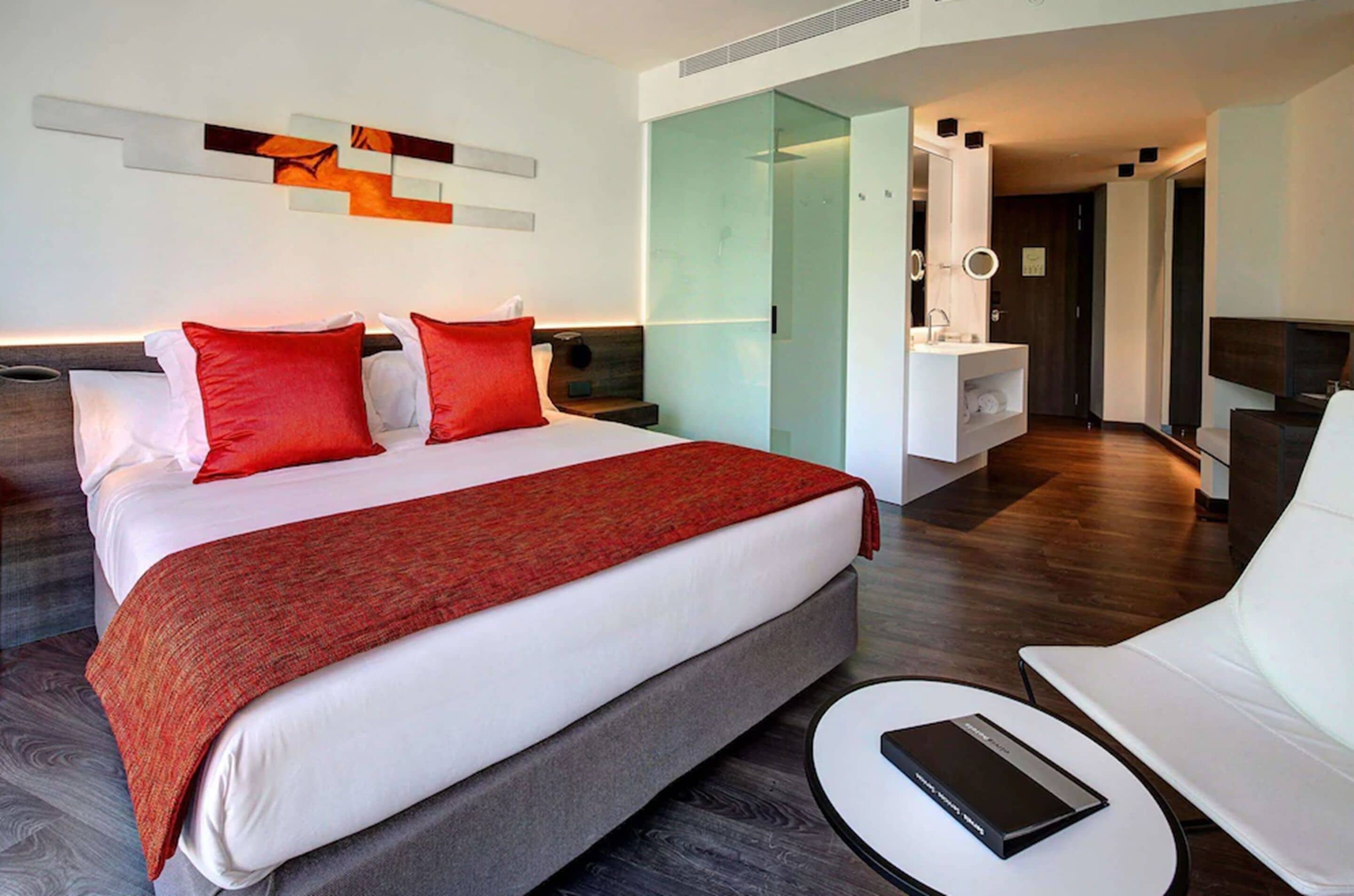 A modern hotel room with a large bed, a sitting area with a white sofa and a small table, a glass-enclosed bathroom, and a minimalist decor.