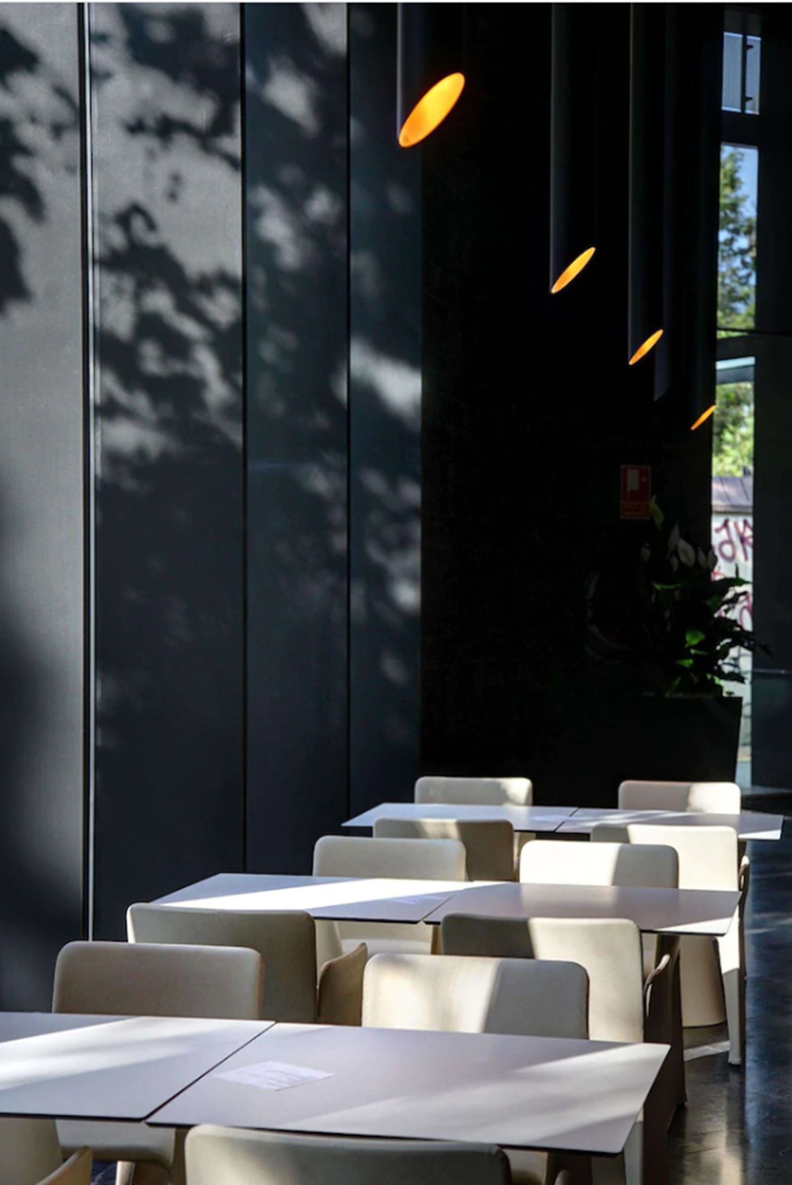 A modern indoor dining area with tables and chairs arranged in rows, large windows allowing natural light, and hanging pendant lights.
