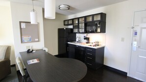A compact kitchen with a dark wooden table, black cabinets, a refrigerator, and a microwave. There is a white door on the right and a bulletin board on the left. - Candlewood Suites Greenville by IHG (Greenville)