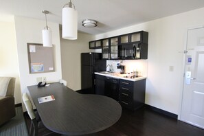 A compact kitchen with a dark wooden table, black cabinets, a refrigerator, and a microwave. There is a white door on the right and a bulletin board on the left. - Candlewood Suites Greenville by IHG (Greenville)