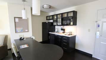 Cocina compacta con cubierta oscura, refrigerador, microondas y lavamanos. Hay una mesa de comedor con sillas y una lámpara colgante.