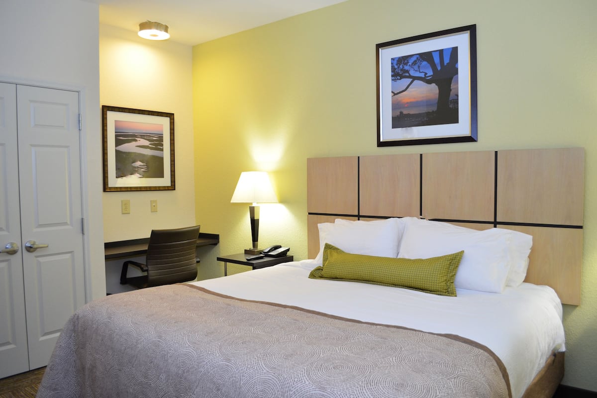 A hotel room with a bed, a desk, a chair, a lamp, and a framed picture on the wall.