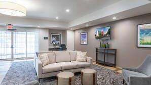 A spacious lobby with a beige sofa, two round ottomans, a gray armchair, a patterned rug, and a television on the wall. - Candlewood Suites Greenville by IHG (Greenville)