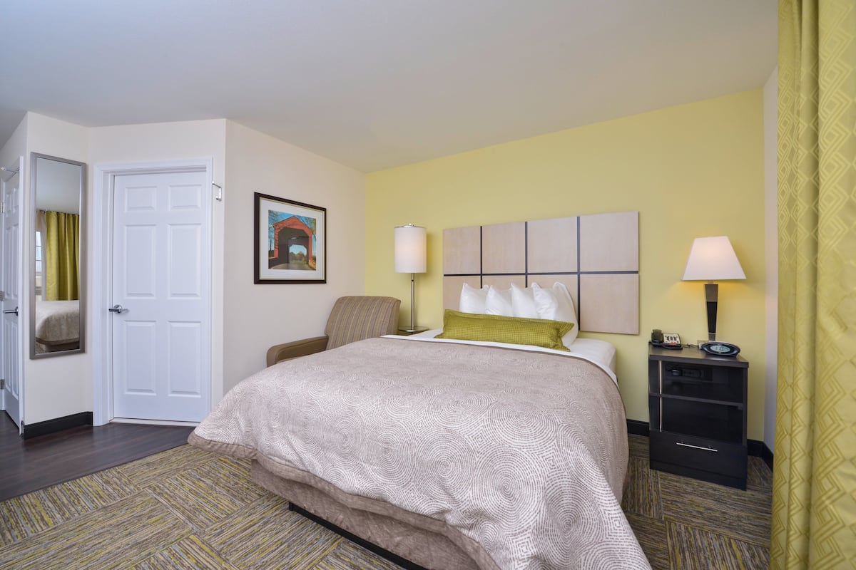 Ein Hotelzimmer mit Bett, Sessel, Nachttisch mit Lampe, gerahmtem Bild an der Wand und Tür.