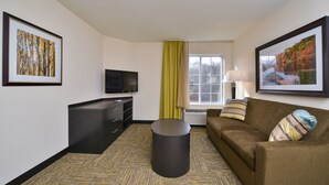 Habitación de hotel con televisión de pantalla plana, sofá, escritorio con cajones, cesto de basura y dos cuadros enmarcados en las paredes.