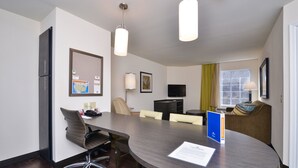 Una habitación de hotel moderna con una mesa de conferencias, sillas de oficina, un televisor de pantalla plana, un refrigerador y una ventana con cortinas.