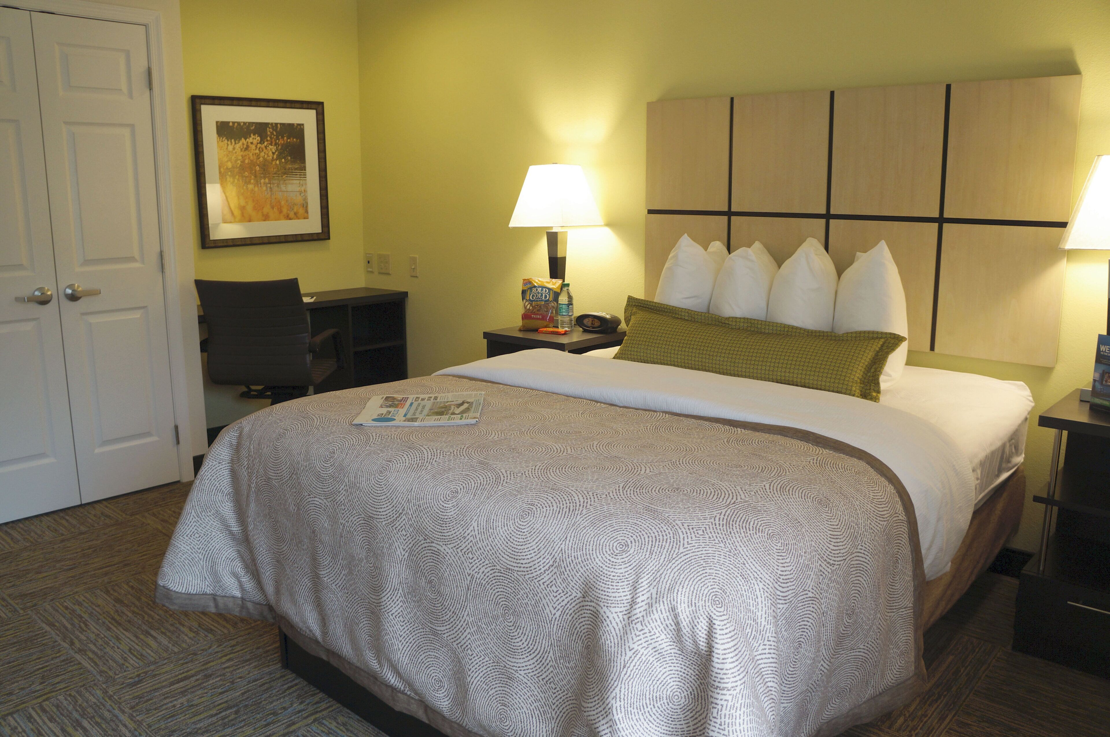 A hotel room with a bed, two bedside lamps, a desk, a chair, a framed picture on the wall, and a door.