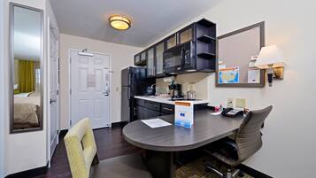 A compact hotel room with a kitchenette, a dining area with a table and chairs, a mirror, and a lamp.