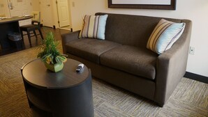A living room with a brown sofa, two striped pillows, a small table with a plant and a remote control, and a framed picture on the wall. - Candlewood Suites Harrisburg - Hershey by IHG (Harrisburg)