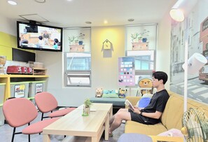 A room with a television, a table with chairs, a sofa, a window with blinds, and a wall clock. - Hostel Tommy (Seoul)