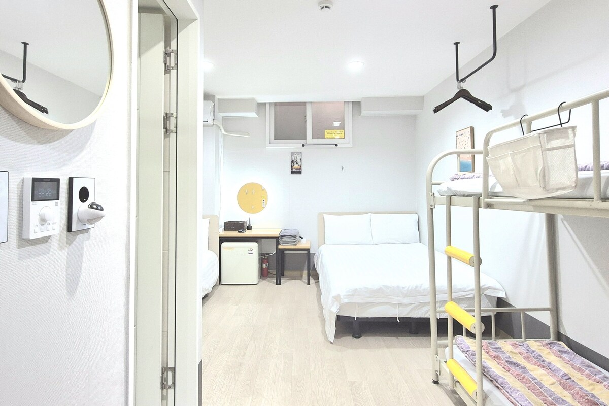 A hotel room with a bunk bed, a desk with a computer, a small fridge, a bedside table, and a mirror.