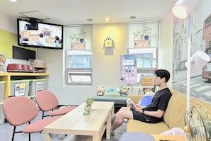 A room with a television, a clock, a window with a view of a building, a sofa, and a table with a plant. - Hostel Tommy (Seoul)