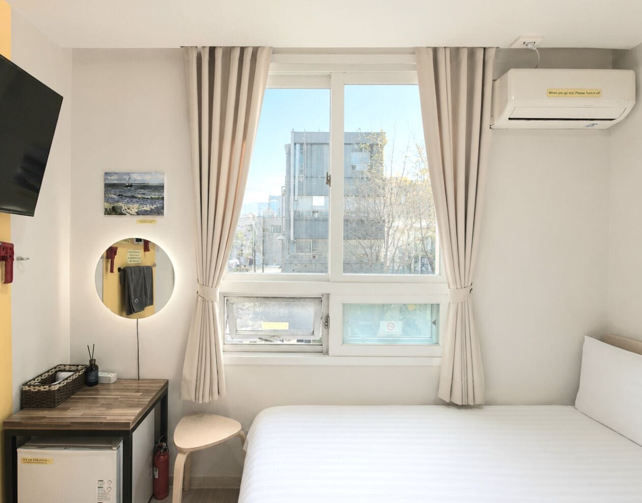 Une chambre d’hôtel comprenant un lit, un bureau avec une lampe, un miroir, une fenêtre avec des rideaux et un appareil de climatisation.