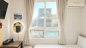 A hotel room with a bed, a desk with a lamp, a mirror, a window with curtains, and an air conditioning unit.