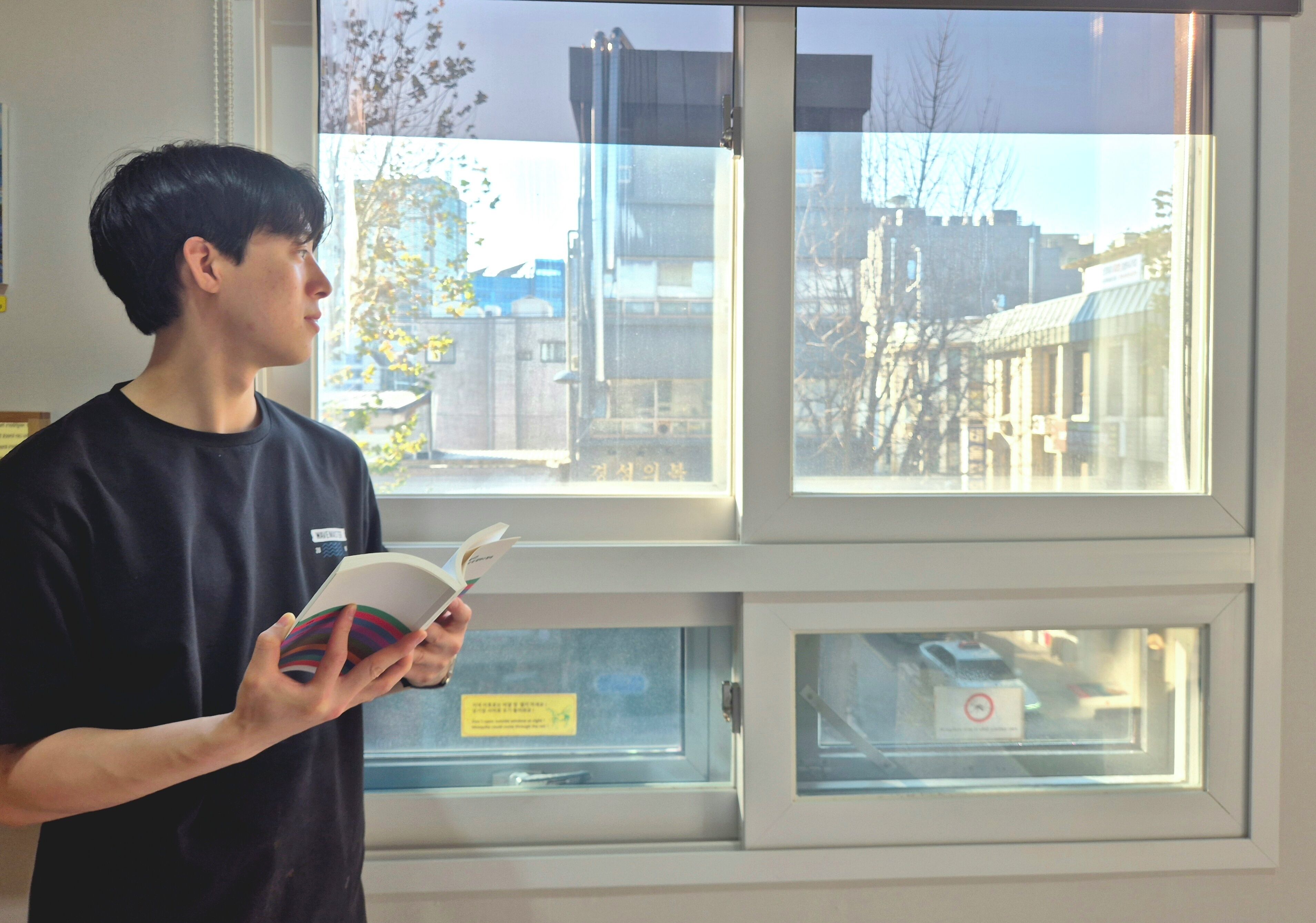 A person reading a book in a room with a window overlooking a cityscape.