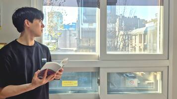 A person reading a book in a room with a window overlooking a cityscape.