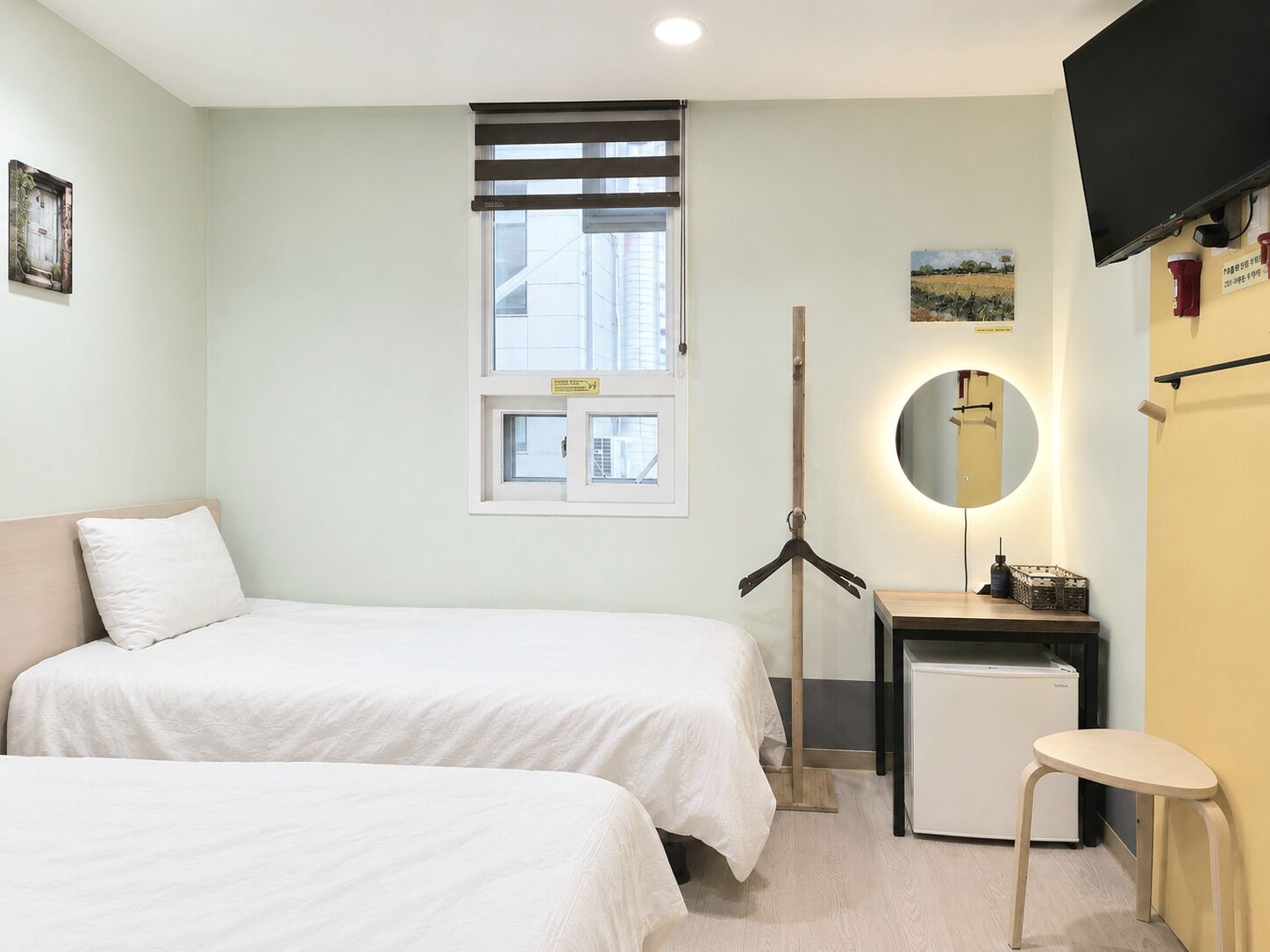 A hotel room with two beds, a small desk with a mirror, a television, and a window with blinds.