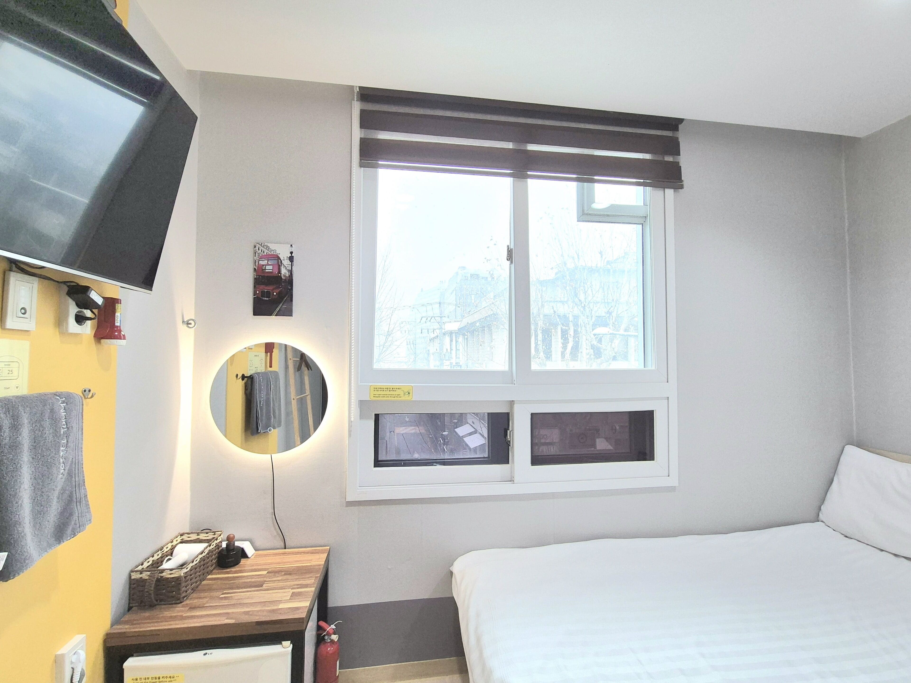Ein Hotelzimmer mit Bett, Nachttisch mit Lampe, einem Wand-Fernseher, einem Fenster mit Jalousien und einem Spiegel.