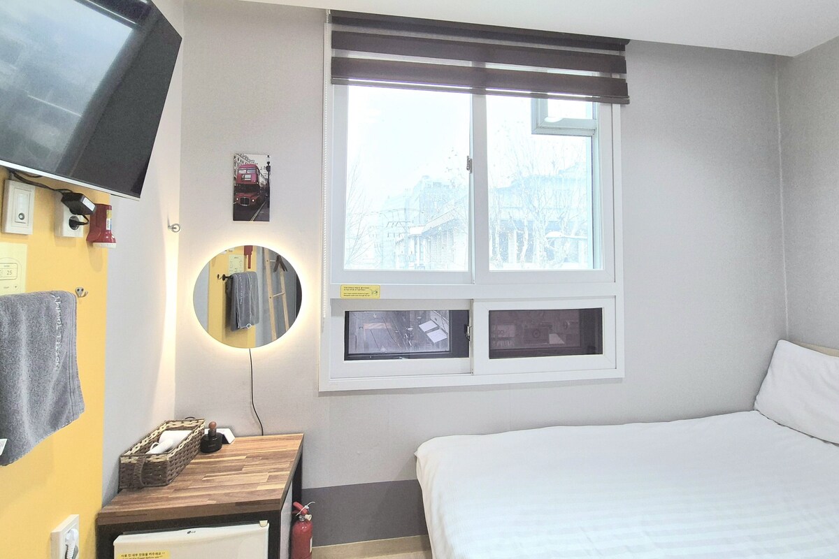A hotel room with a bed, a bedside table with a lamp, a mounted TV, a window with blinds, and a mirror.