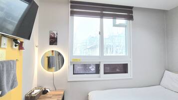 Ein Hotelzimmer mit Bett, Nachttisch mit Lampe, einem Wand-Fernseher, einem Fenster mit Jalousien und einem Spiegel.