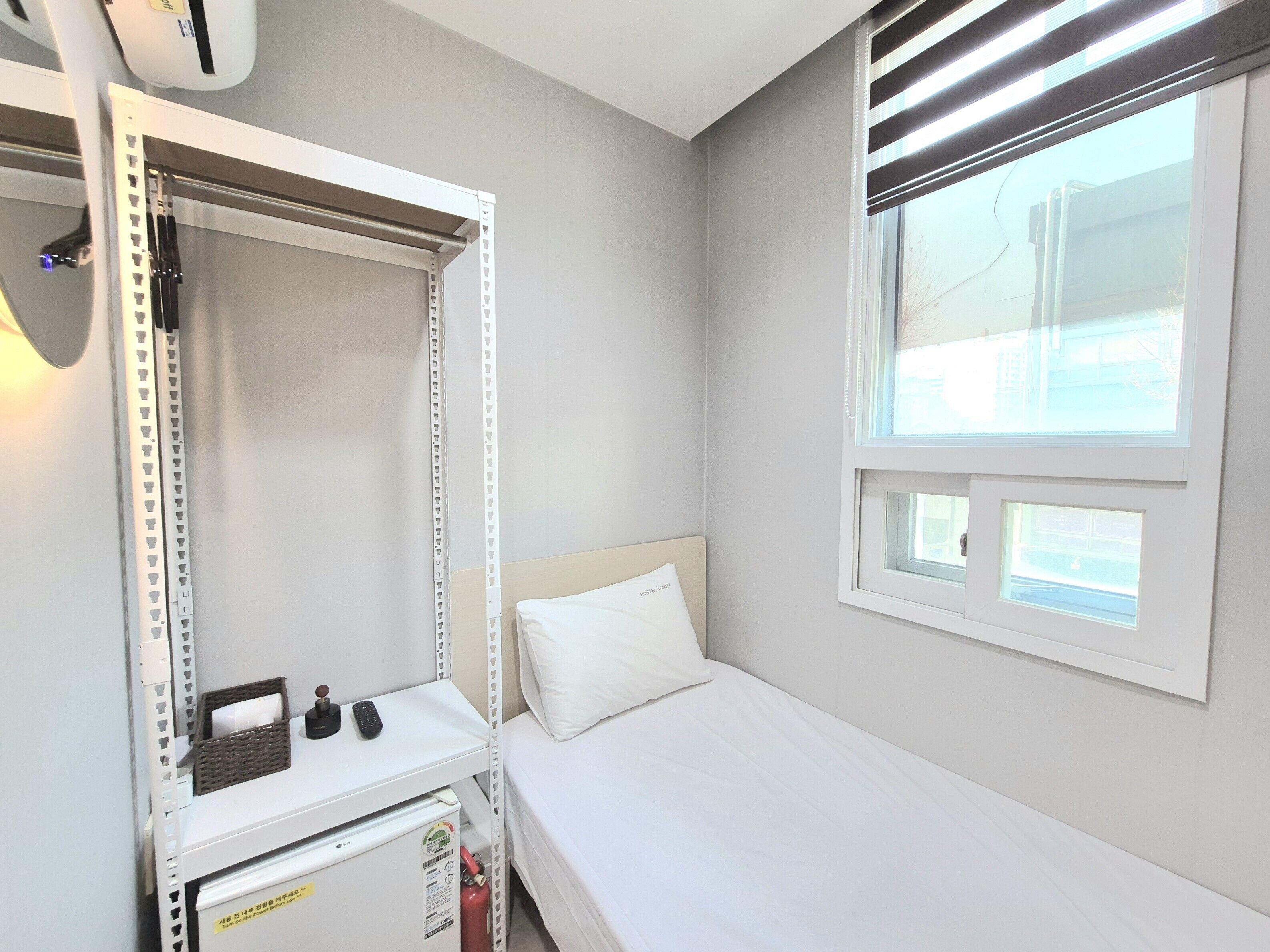 Una cama individual con cabecera blanca, una mesita de noche pequeña con un control remoto, una pared blanca y una ventana con persianas.