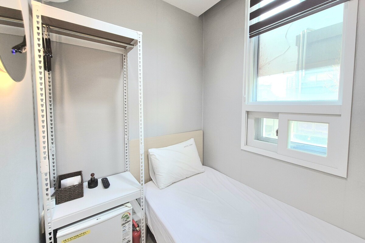 A single bed with a white headboard, a small bedside table with a remote control, a white wall, and a window with blinds.